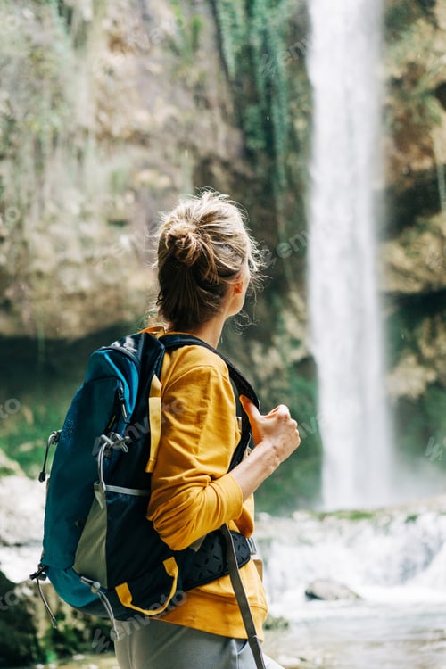 Preview: Young active unrecognizable woman from behind with a backpack looking at a waterfall in a canyon.
