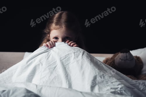 Preview: scared child hiding under blanket while sitting on bedding near teddy bear isolated on black