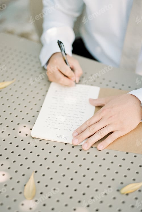 Preview: Groom writes an oath with a pen on a piece of paper while sitting at the table. Cropped. Faceless