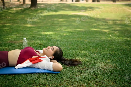 Preview: Carefree young woman listening to music while resting on yoga mat in a sunny park