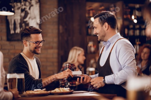 Preview: Young happy man making mobile payment in a pub.
