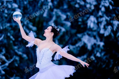 Preview: Ballerina in white tutu joyfully dances with a glass sphere in a snowy forest setting. WA, USA