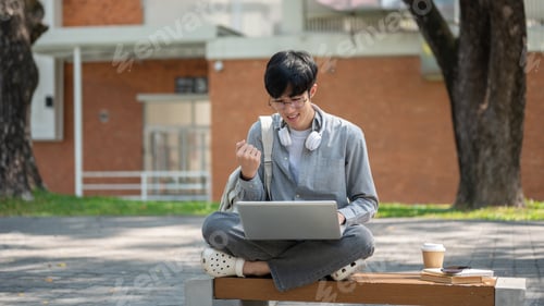 Preview: An asian man with glasses is making a fist while looking at his laptop, sitting on sidewalk bench.