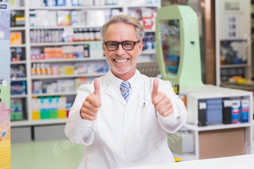 Preview: Senior male pharmacist wearing lab coat giving thumbs up behind counter with medicine shelves