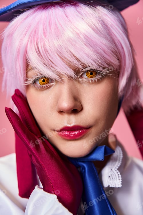 Preview: good looking chic female cosplayer in blue hat and vivid dress looking at camera on pink backdrop