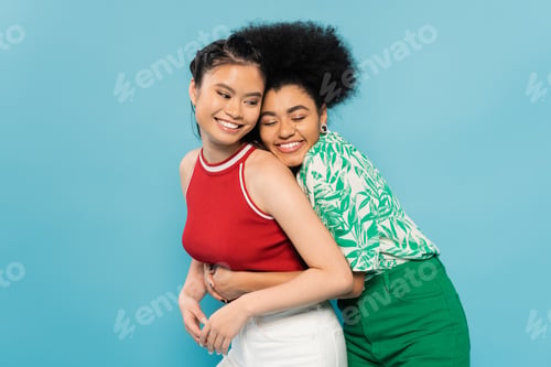 Preview: happy african american woman with closed eyes embracing smiling asian friend isolated on blue