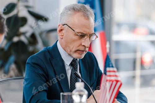Preview: selective focus of bearded diplomat in eyeglasses near american flags