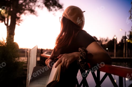 Preview: Young girl reading a book on a bridge at sunset in a park with trees
