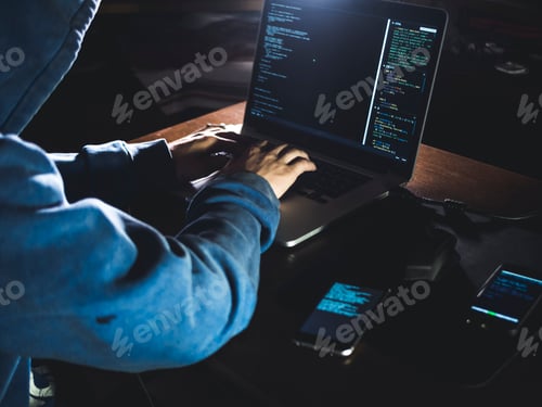 Preview: anonymous person in the hood sitting in front of computer working with laptop and a mobile phone