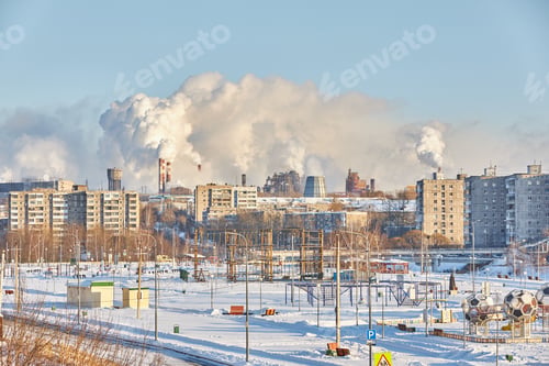 Preview: Buildings stand in a city covered by snow. Smoke billows from factories in the background