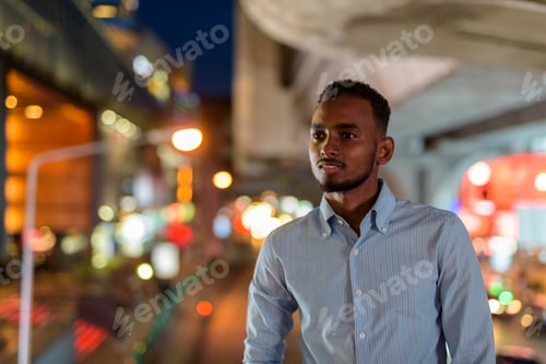 Preview: Portrait of handsome black African businessman outdoors in city at night smiling and thinking