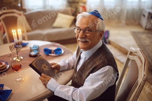 Preview: Happy mature Jew reading Hebrew bible at home and looking at camera.