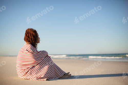 Preview: Side view of beautiful mixed race woman wrapped in blanket sitting on beach in the sunshine