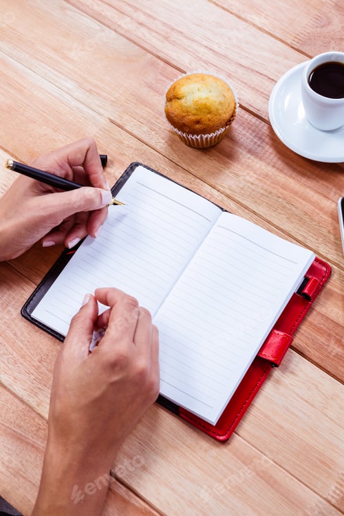 Preview: Overhead of feminine hands writing on agenda with espresso, smartphone and muffin on table