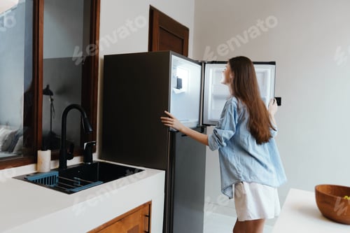 Preview: A young woman happily exploring her fridge in a modern kitchen, dressed casually in a denim shirt
