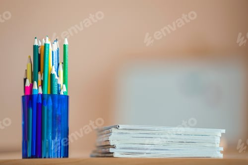 Preview: Wooden colorful drawing pencils arranged in plastic jug and stack of copybooks on light copy space