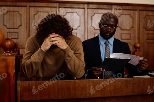 Preview: Young upset male suspect with clasped hands by his head sitting next to attorney