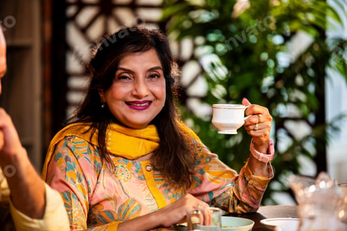 Preview: Elderly Indian lady enjoys tea at home while smiling and holding a cup at dining table