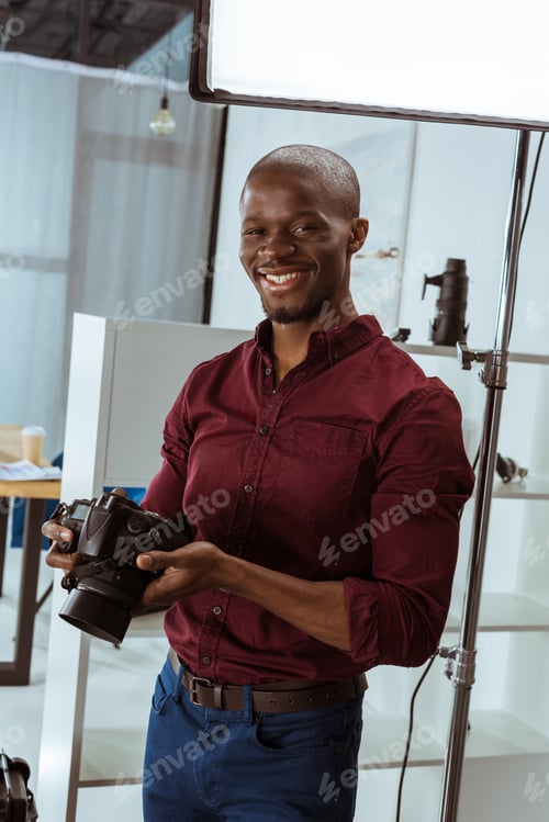 Preview: portrait of cheerful african american photographer with photo camera in hands looking at camera in
