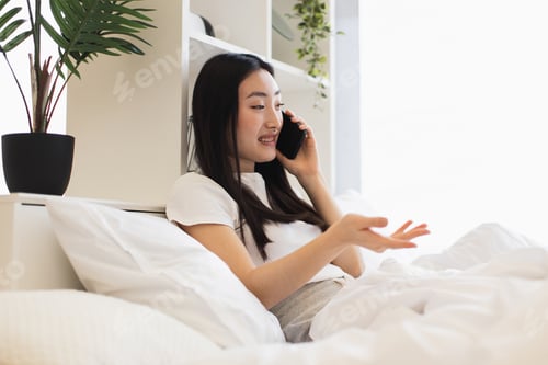 Preview: Asian woman enjoying a phone conversation while relaxing in bed at home