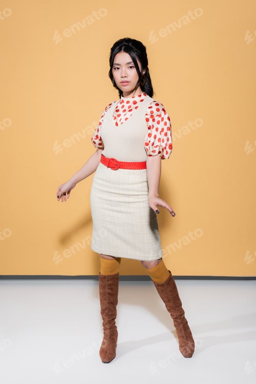 Preview: Full length of trendy asian model in retro clothes looking at camera on orange background