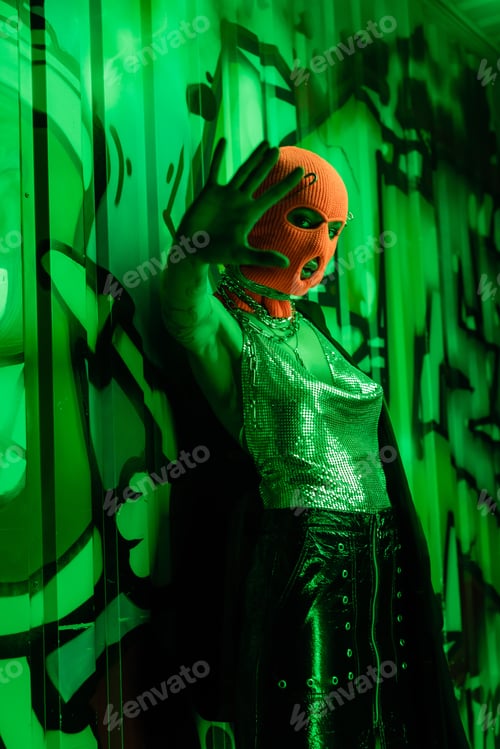 Preview: sexy woman in balaclava and silver top standing with outstretched hand near wall with graffiti in