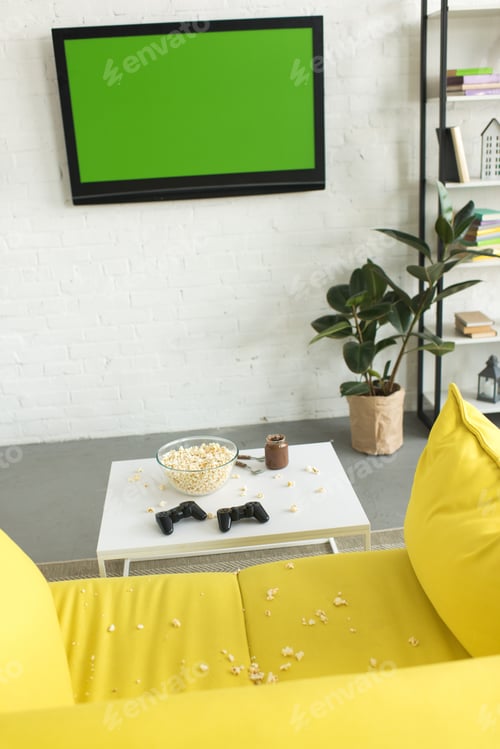 Preview: high angle view of glass bowl with popcorn and joysticks on table near yellow sofa in living room