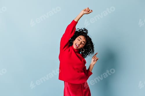Preview: Stylish mixed race girl posing with closed eyes. Blissful curly woman in red clothes dancing on blu
