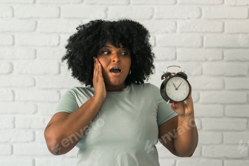 Preview: Surprised African American woman holding an alarm clock while expressing shock against a textured