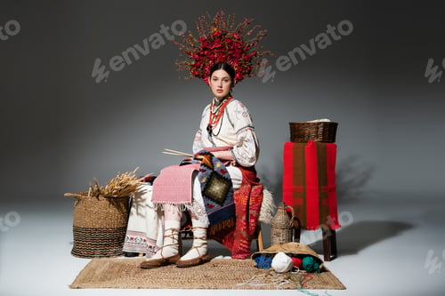 Preview: full length of young ukrainian woman in traditional clothes and red floral wreath holding knitting