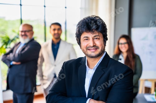 Preview: Confident Indian business leader standing with folded hands, colleagues out of focus in the office