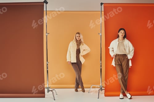 Preview: fall fashion, young interracial women in stylish attire posing on pastel multicolored backdrop