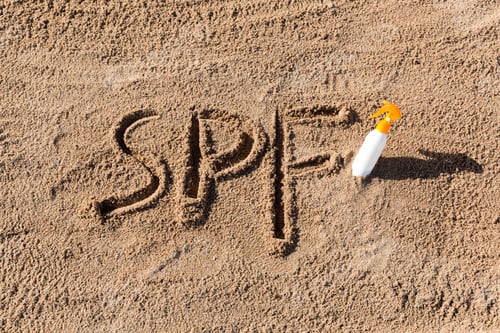 Preview: Sun protect factor. SPF word written on the sand and white bottle with suntan cream.