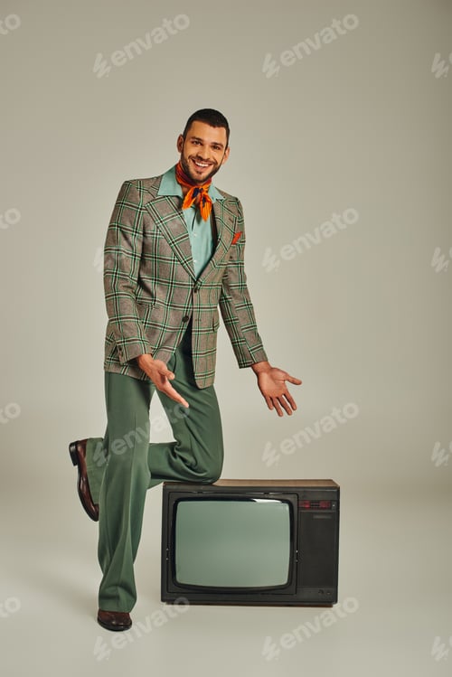 Preview: smiling man in elegant retro style attire pointing at vintage tv set on grey backdrop, full length