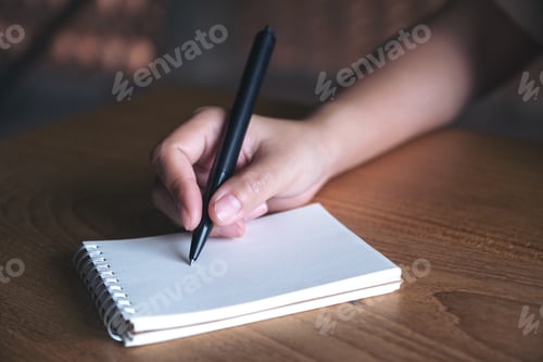 Preview: Closeup image of a hand writing down on a white blank notebook on wooden table