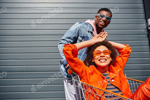 Preview: Cheerful young african american woman in sunglasses and bright outfit sitting in shopping cart