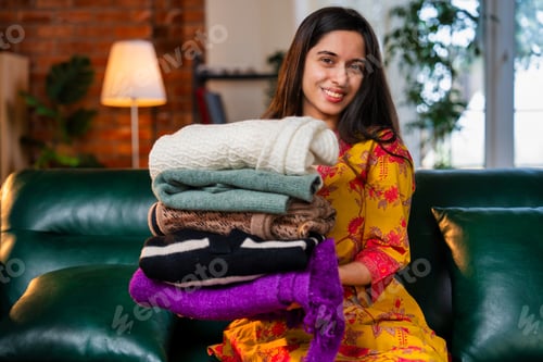 Preview: Indian woman in kurta folding woolen clothes, enjoying the fresh fragrance while relaxing on sofa