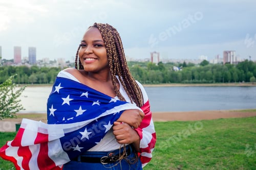 Preview: Smiling afro american woman holding USA flag and looking at camera autumn evening