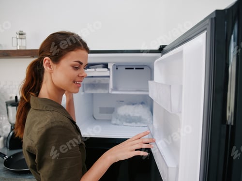 Preview: Woman smiling with teeth looking into camera in kitchen at home opened freezer empty with ice inside