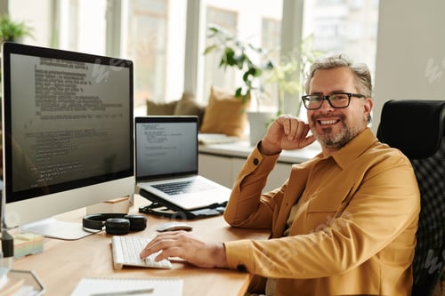 Preview: Successful mature IT engineer looking at camera by workplace
