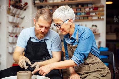 Preview: Senior woman spinning clay on a wheel with teacher at pottery class