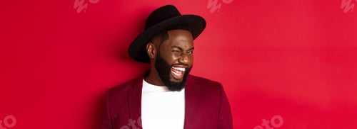 Preview: Cheeky african american man in party outfit, winking at camera and smiling, standing against red