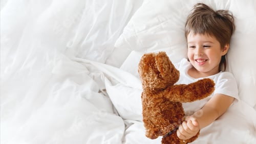 Preview: Toddler lies in bed with cute teddy bear. Little boy under white blanket with fluffy toy.