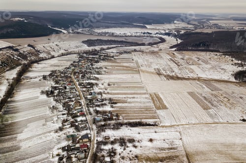 Preview: Top view of empty snowy fields, houses along road and woody hills on blue sky background. Aerial