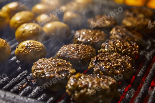 Preview: Closeup of kebabs with grilled potatoes in the background and smoke from the barbecue. BBQ party
