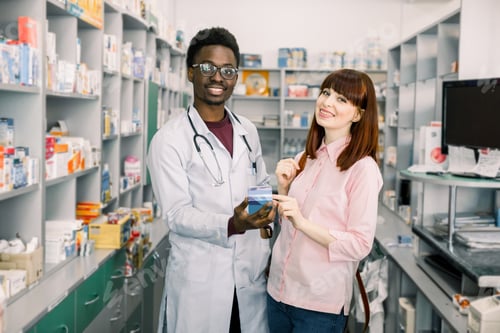 Preview: Happy young woman customer standing near African man pharmacist, holding medicine together