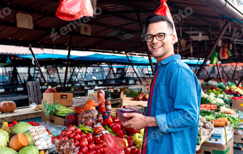 Preview: Portrait of young man standing in front of a vegetable counter in an outdoor market.