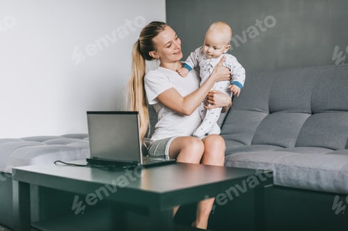 Preview: happy young mother holding adorable infant child while using laptop at home