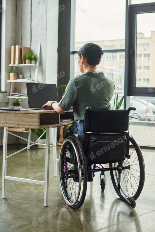 Preview: dedicated brunette disabled woman in casual attire sitting in wheelchair and working hard in office