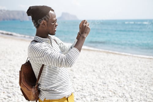 Preview: Side view of African American young man with backpack, in hat and striped shirt taking photos of sea
